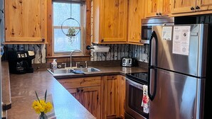 A kitchen with wooden cabinets, stainless steel appliances, a central island with a vase of flowers, and a window with a view of trees. - Pine Lane is your cozy, pet-friendly escape -w/ hot tub, fire pit, & peaceful vibes near Mohican (Butler)