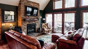 A cozy living room with a stone fireplace, leather sofas, and large windows. - Pine Lane is your cozy, pet-friendly escape -w/ hot tub, fire pit, & peaceful vibes near Mohican (Butler)