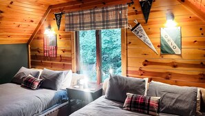 Een gezellig cabin-kamer met twee bedden, een raam met ruitjesgordijnen en decoraties aan de muur, waaronder een bord met de tekst 'Keep it Wild' en een spandoek met 'Life is Better in the Woods'.