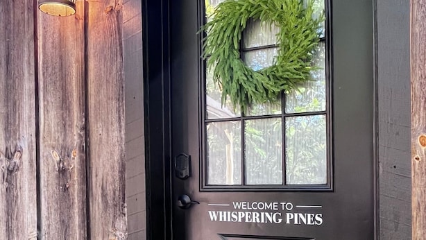 En sort dør pyntet med en krans og teksten "WELCOME TO WHISPERING PINES".