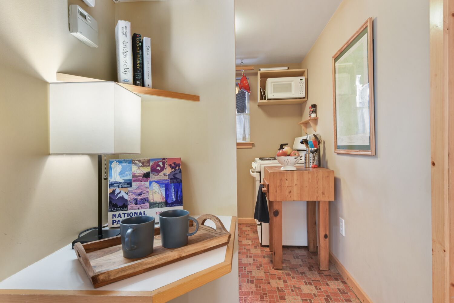 A compact kitchen with a microwave, a stove, and a small sink. There are books on a shelf and a tray with mugs on a table.