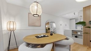 Un espace repas moderne avec une table ronde en bois, deux chaises grises à revêtement rembourré, un luminaire suspendu, une œuvre d’art encadrée et un miroir au mur.