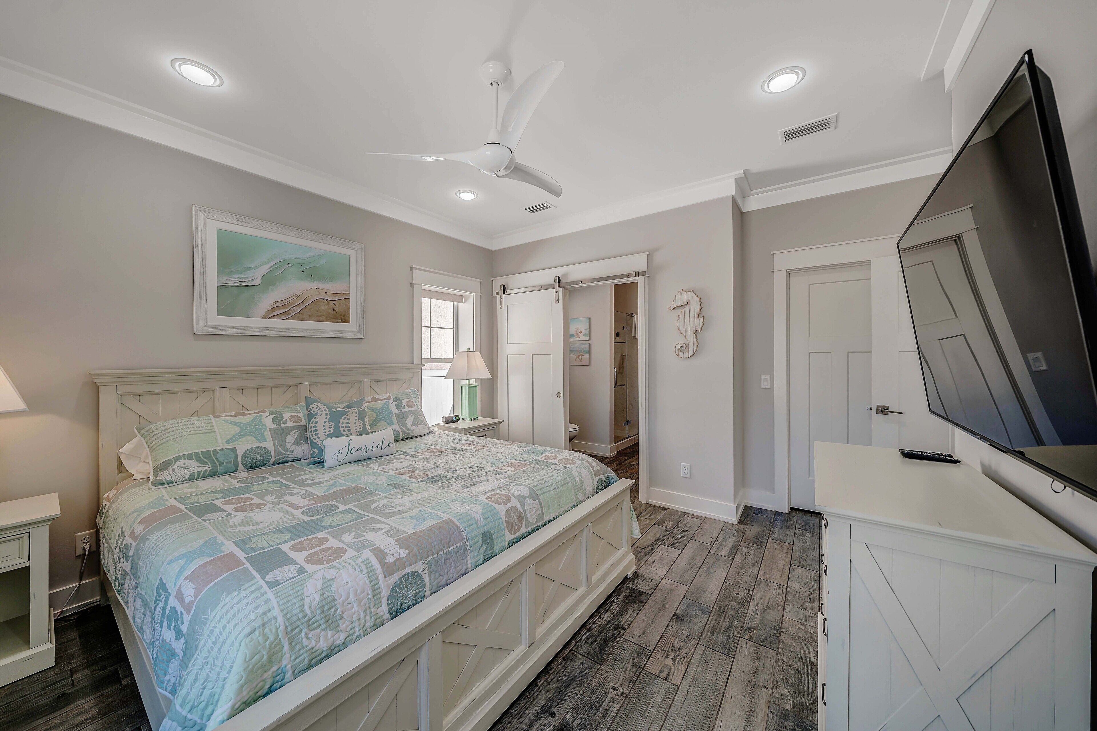 A bedroom with a bed, a ceiling fan, a TV, and a door leading to a bathroom.