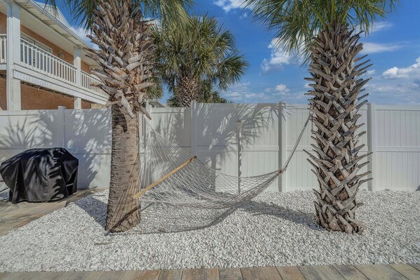 A hammock between two palm trees, a white fence, and a building with a balcony in the background.