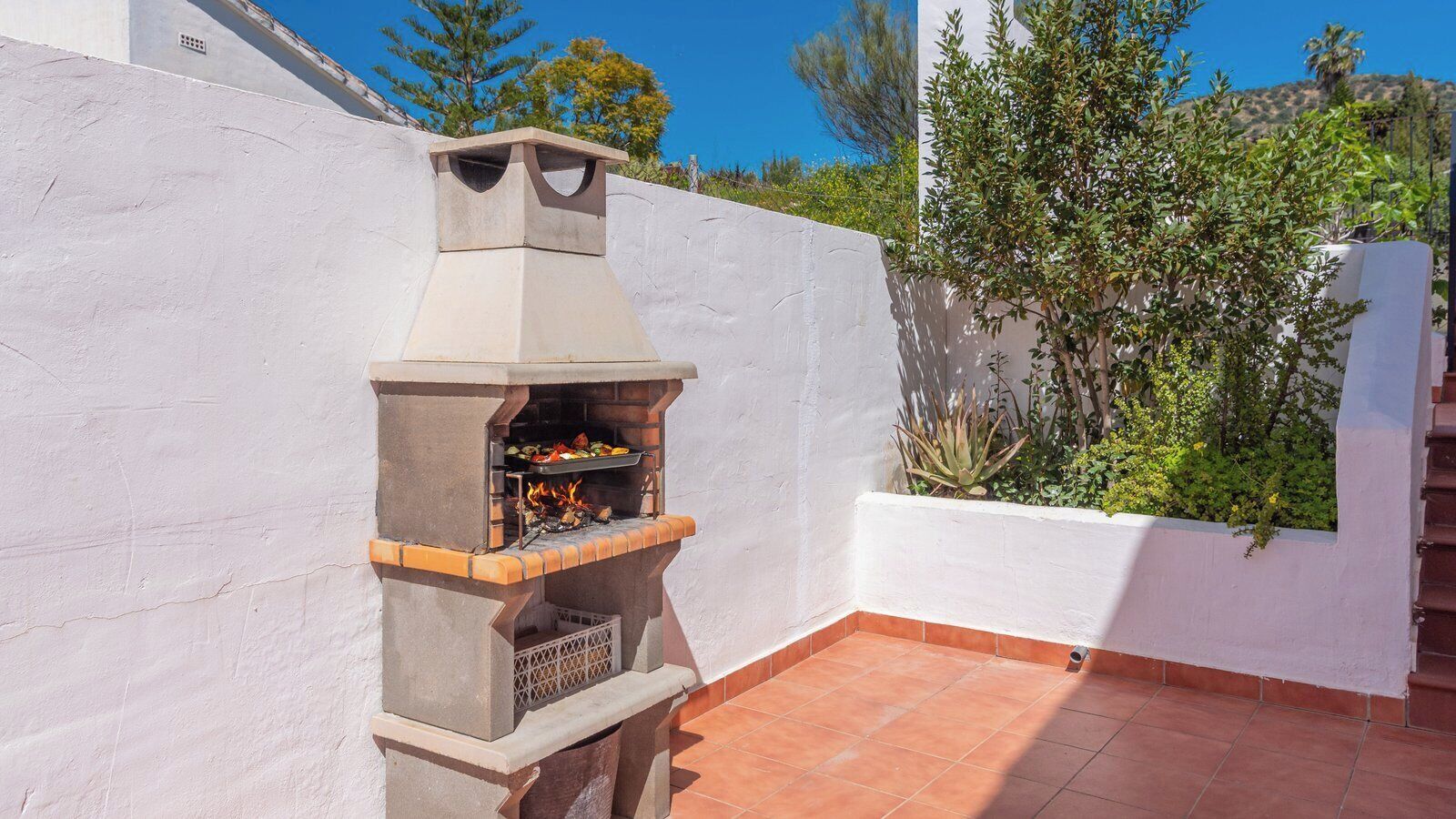 Ein aus Stein gemauerter Außenofen mit Schornstein, umgeben von weißen Wänden und einer gefliesten Terrasse.