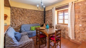 Una sala acogedora con pared de piedra, un sofá azul con cojines estampados, una mesa de comedor de madera con sillas y una ventana con cortinas.