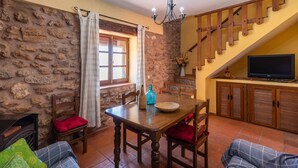 Un comedor acogedor con pared de piedra, mesa de madera y sillas. Un televisor está montado en la pared y se ve una escalera al fondo.