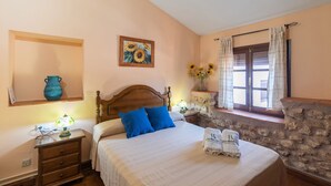 Un dormitorio con cama de madera, una mesita de noche con lámpara, pared de piedra y ventana con cortinas.