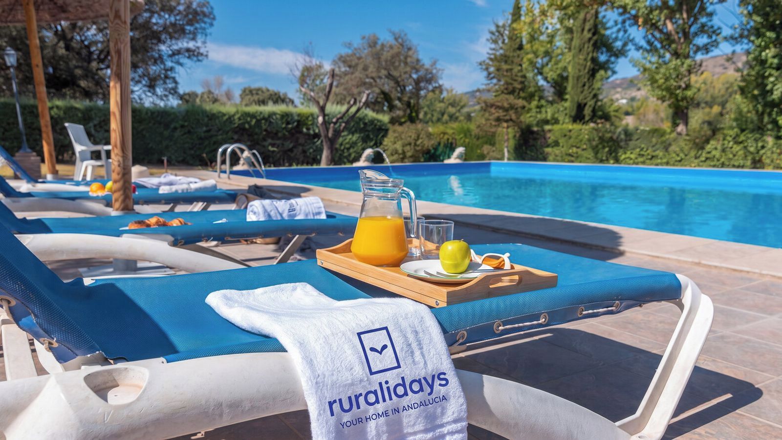 Ein Poolbereich mit Liegestühlen, einem Tablett mit Orangensaft und einem Handtuch mit dem Logo 'ruraldays'.