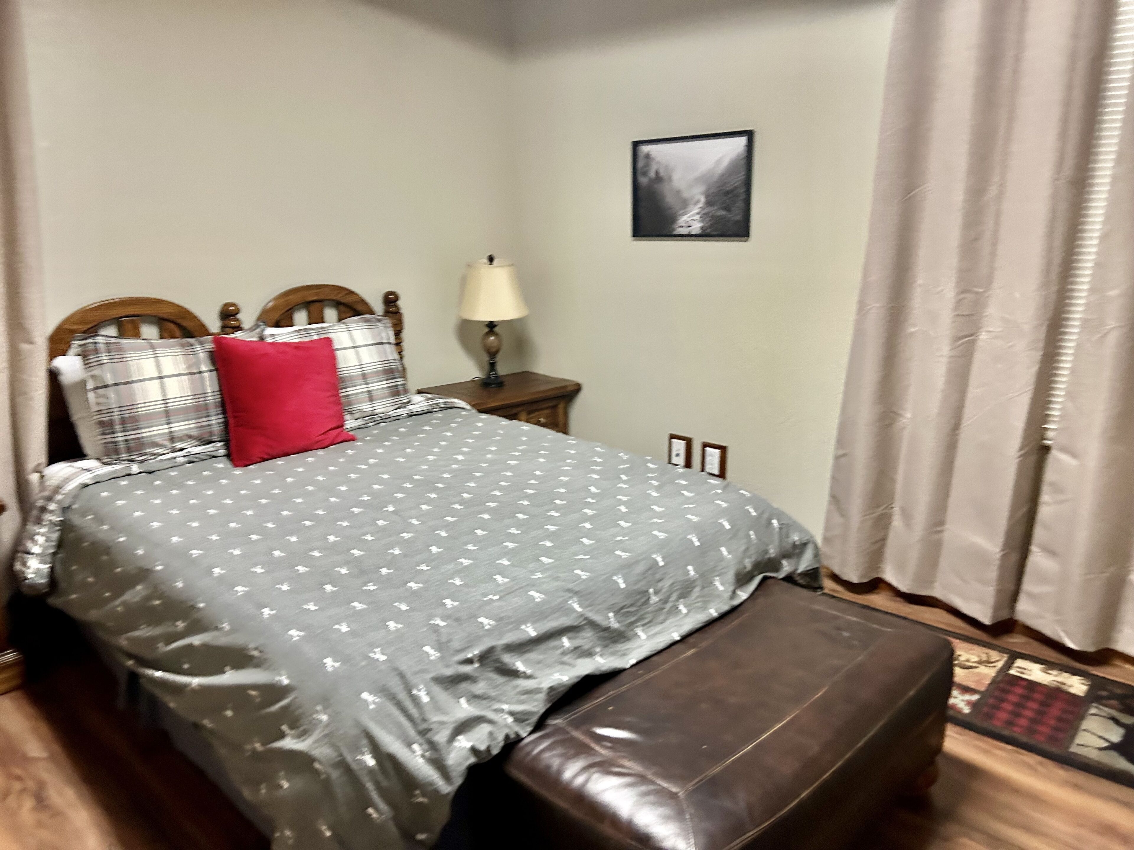 Un lit bien fait, recouvert d’une couvre-lit grise à motifs, avec un oreiller rouge et deux têtes de lit en bois.