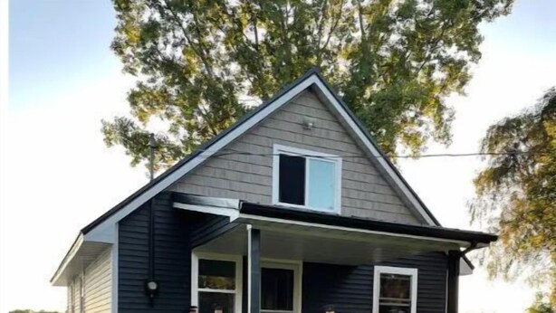 A two-story house with a dark exterior, a front porch with a wooden railing, and a stone pathway leading to the entrance. - Whispering Pines Lake House (Gladwin)