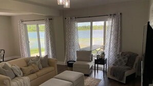 A spacious living room with a large sofa, ottoman, and a smaller chair. There are two windows with curtains, a small table with a plant, and a view of a body of water. - Whispering Pines Lake House (Gladwin)