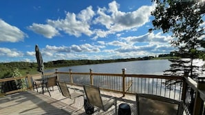 A wooden deck with chairs and a table overlooking a lake, with a railing and trees in the background. - Whispering Pines Lake House (Gladwin)