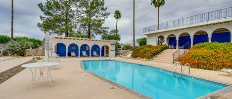 Una piscina con borde de azulejos azules, un área de descanso techada con puertas azules y un paseo bordeado de palmeras.