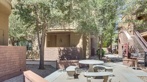 Un espace extérieur aménagé en terrasse, avec une table et des bancs en pierre, entouré d’arbres et bordé d’un bâtiment doté d’un escalier.