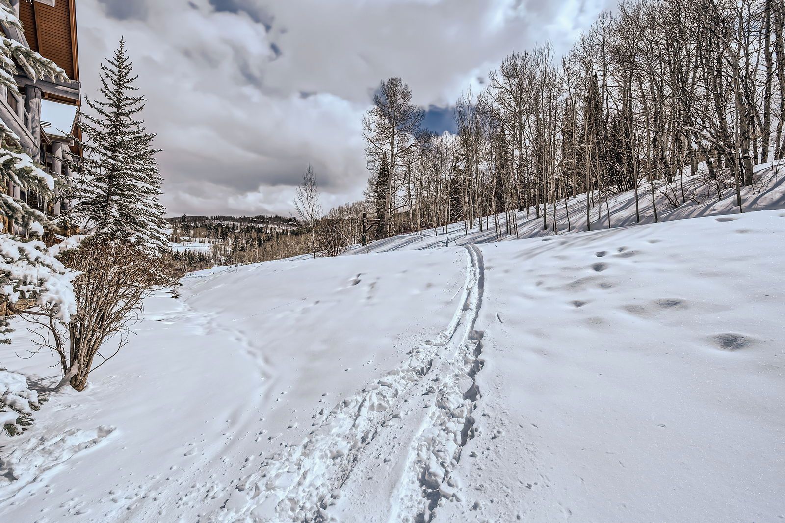 A snow-covered ski trail with footprints, a wooden building on the left, and leafless trees.
