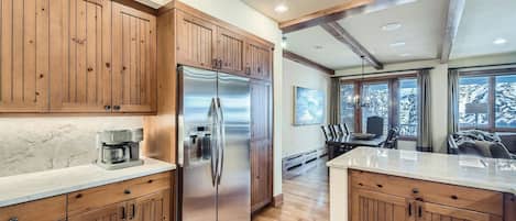 Una cocina con armarios de madera, un refrigerador de acero inoxidable, una cafetera y un comedor con vista a árboles cubiertos de nieve.