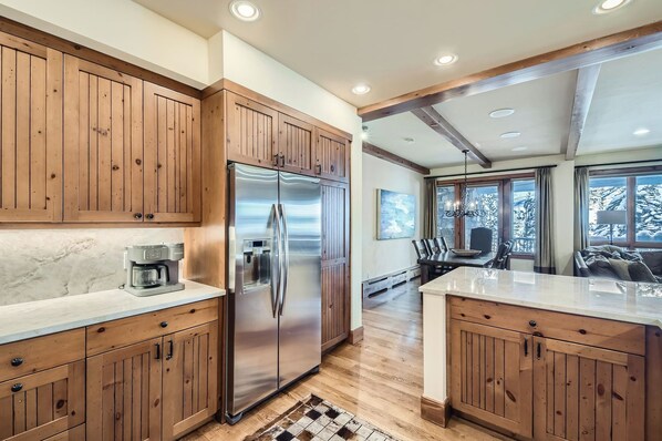 A kitchen with wooden cabinets, a stainless steel refrigerator, a coffee maker, and a dining area with a view of snow-covered trees. - Charlie's Slopeside: Bear Paw, Ski-In/Out, Sleeps 8, Bachelor Gulch + Ritz Pool & Hot Tub Access (Beaver Creek)