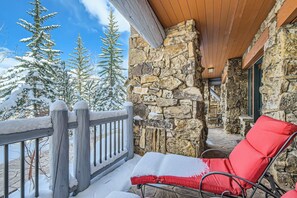 Un muro exterior de piedra, una terraza de madera y una silla roja con un cojín cubierto de nieve.
