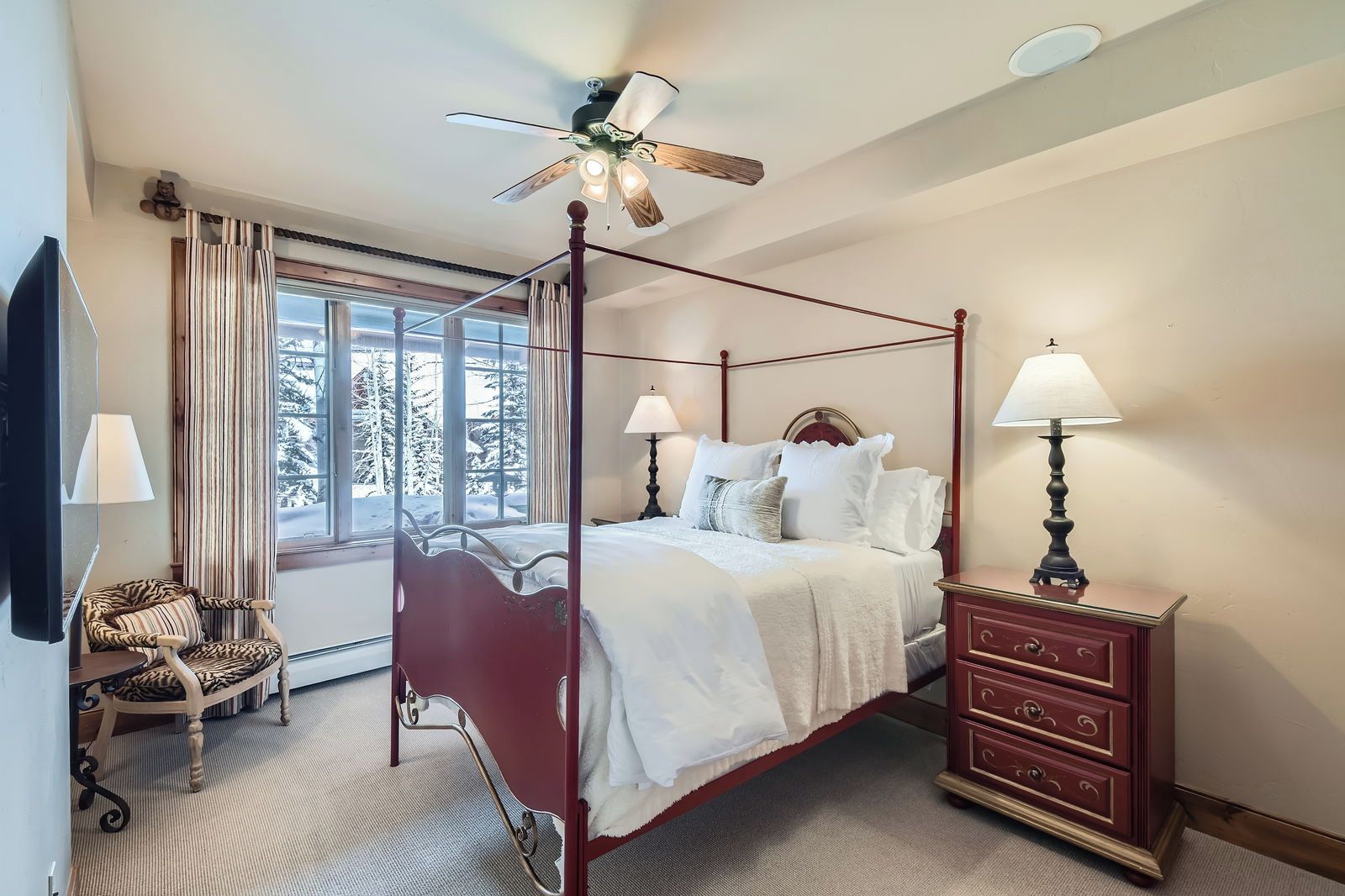 A four-poster bed with white bedding, a wooden nightstand with a lamp, a chair with striped upholstery, and a window with a view of snow-covered trees.