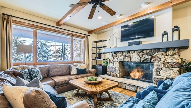 A cozy living room with a stone fireplace, leather sofas, a wooden coffee table, and a large window with a view of snow-covered trees. - Spacious 3BR ski-in/out Beaver Creek condo with valley views. (Beaver Creek)