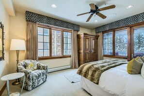 Une chambre avec un grand lit, un ventilateur de plafond, une armoire en bois et une fenêtre donnant sur les arbres.