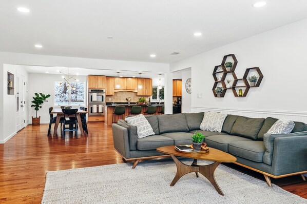 A modern living room with a grey sectional sofa, wooden coffee table, and a dining area with a wooden table and chairs. - Birch House | Sauna, Nursery + Pet-Friendly | Private Grand Traverse Bay Access (Suttons Bay)