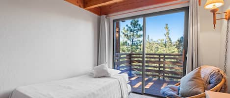 A bedroom with a single bed, a wicker chair, a wooden ceiling, and a large sliding glass door leading to an outdoor deck.