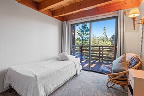 A bedroom with a single bed, a wicker chair, a wooden ceiling, and a large sliding glass door leading to an outdoor deck. - TreeHouse on Rainbow (Big Bear City)
