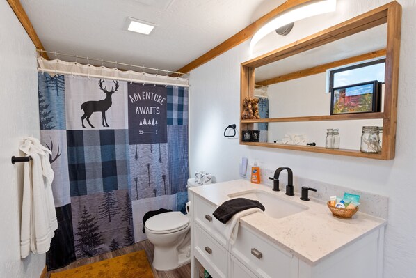 A bathroom with a shower curtain featuring a deer and mountains, a white toilet, a sink with a mirror, and a shelf with toiletries.