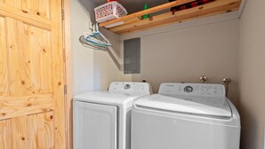 A laundry room with two white front-loading washing machines, wooden cabinets, and a ceiling-mounted shelf with a laundry basket and a green bottle. - TreeHouse on Rainbow (Big Bear City)