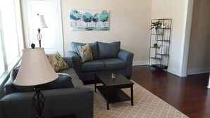 Un salon avec un canapé gris, un fauteuil à motifs, une table basse, un lampadaire, un tableau représentant des arbres et un présentoir à plantes en métal.