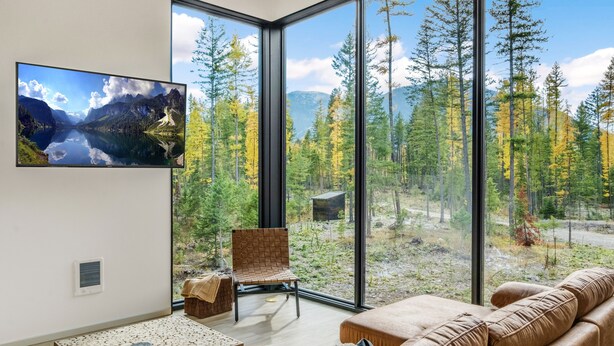 A modern living room with a large window offering a view of a forest and mountains. - Off Grid Stays - Treehouse #3 (Columbia Falls)