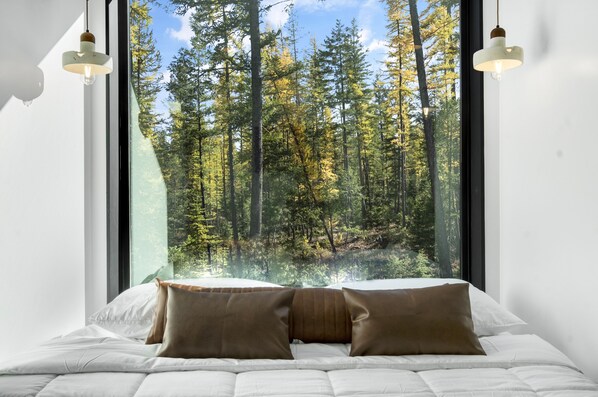 A bedroom with a large window overlooking a forest. There are two bedside lamps and two pillows on the bed.