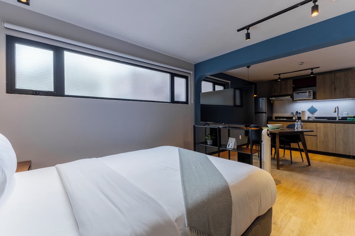 A modern hotel room with a large bed, a kitchenette, and a dining area.