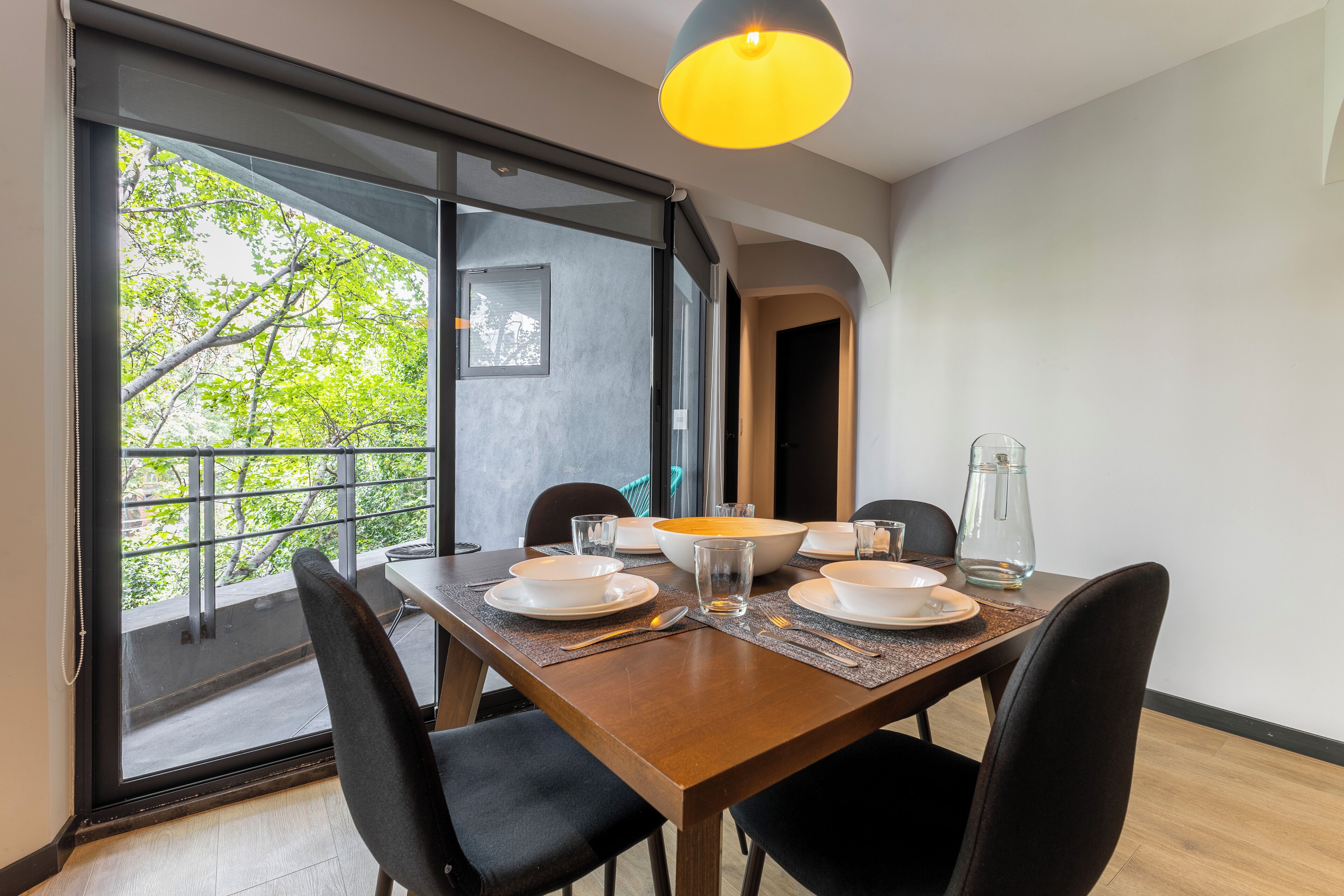 A dining area with a wooden table set for four, large windows, and a view of greenery outside.