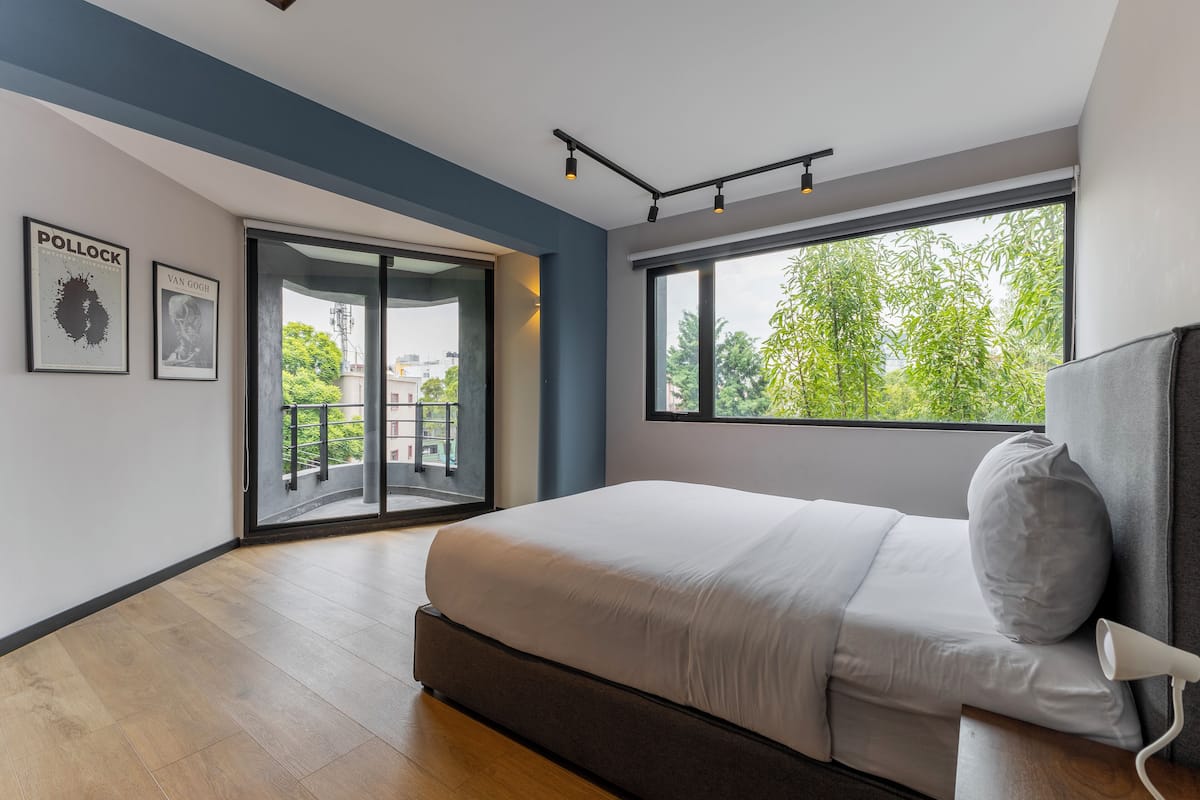 A modern bedroom with a large bed, a sliding glass door to a balcony, and a large window offering a view of greenery.