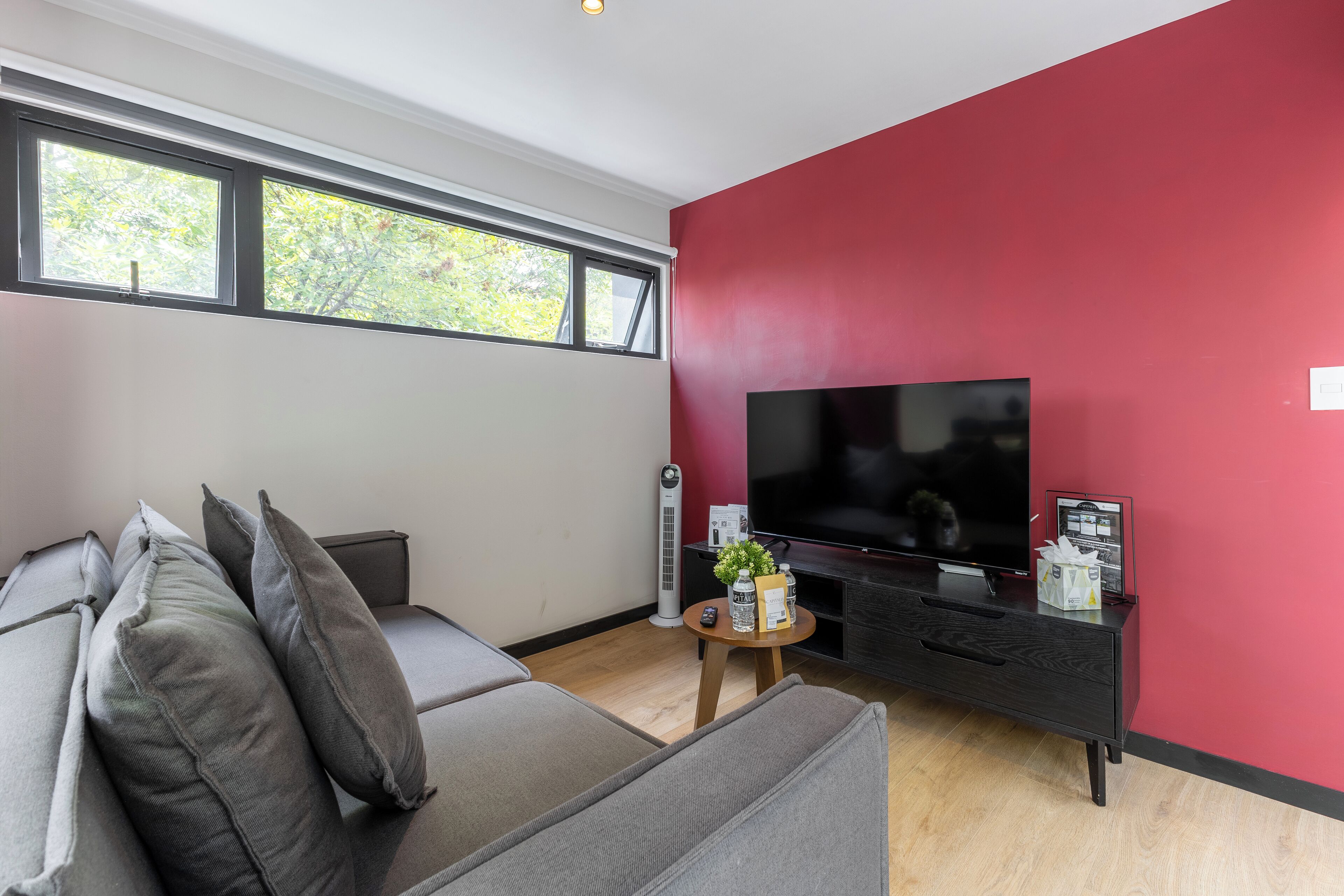 Una sala de estar moderna con un sofá gris, un televisor de pantalla plana sobre un soporte negro, una pequeña mesa de madera con una planta y una pared de acento roja.