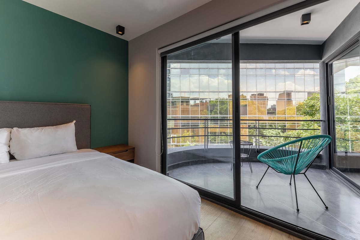 A modern hotel room with a large bed, a balcony with a chair, and a city view.