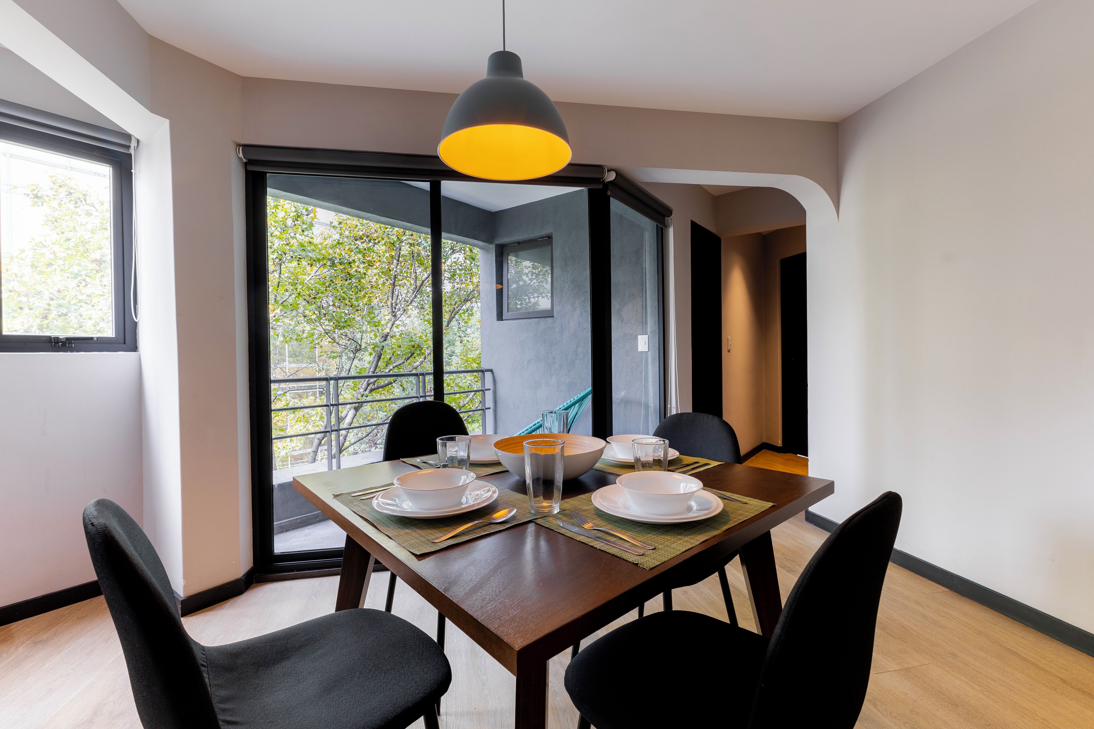 A dining area with a table set for four, a large window with a view of trees, and a hanging pendant light.