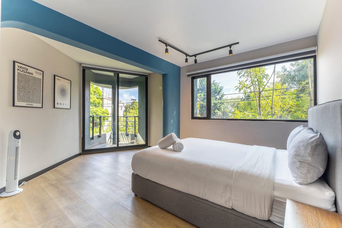 A modern bedroom with a large bed, a ceiling fan, and a view of greenery outside.