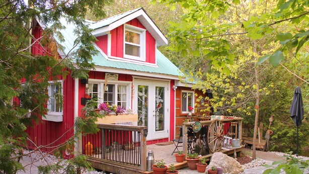A red wooden house with a green roof, surrounded by potted plants and a stone pathway. - Gorgeous cozy cottage besides a bustling stream, surrounded by gorgeous greenery (Orangeville)