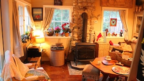 A cozy living room with a stone wall, a fireplace, a comfortable chair, a table with a teapot, and a window with curtains. - Gorgeous cozy cottage besides a bustling stream, surrounded by gorgeous greenery (Orangeville)