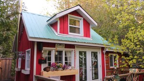 A red cottage with a green roof, a front porch with a bench, and a sign that reads "Enchanted Cottage." - Gorgeous cozy cottage besides a bustling stream, surrounded by gorgeous greenery (Orangeville)