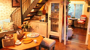 A cozy living room with a wooden staircase, a dining table with a floral tablecloth, and a comfortable seating area. - Gorgeous cozy cottage besides a bustling stream, surrounded by gorgeous greenery (Orangeville)