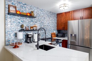 Una cocina con encimera de mármol blanco, refrigerador de acero inoxidable, estufa negra y gabinetes de madera.