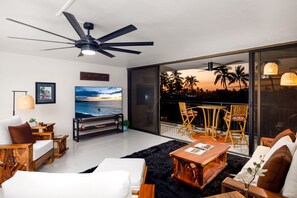 Una sala moderna con un televisor grande, muebles de madera, un ventilador de techo y vistas a palmeras y al atardecer a través de amplios ventanales.
