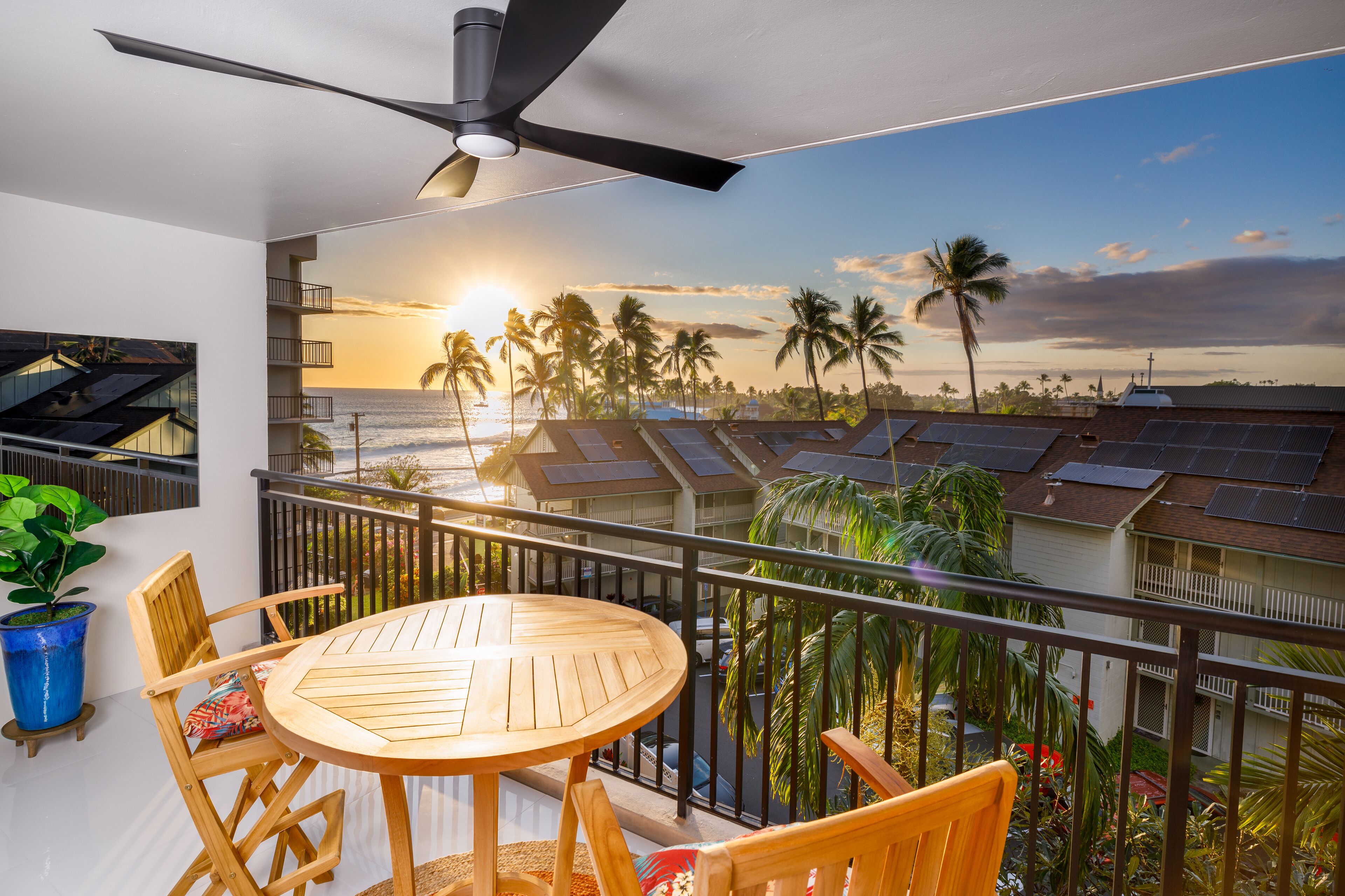A balcony with a table and chairs, overlooking a sunset, palm trees, and solar panels.