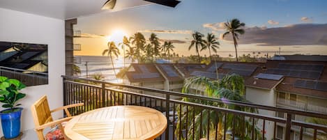 A balcony with a table and chairs, overlooking a sunset, palm trees, and solar panels.