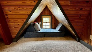 Une chambre mansardée chaleureuse, avec un plafond en pente, une petite fenêtre et un coin salon intégré avec des coussins.